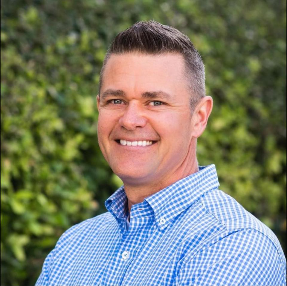 Man smiling outdoors, wearing blue checkered shirt, greenery background.